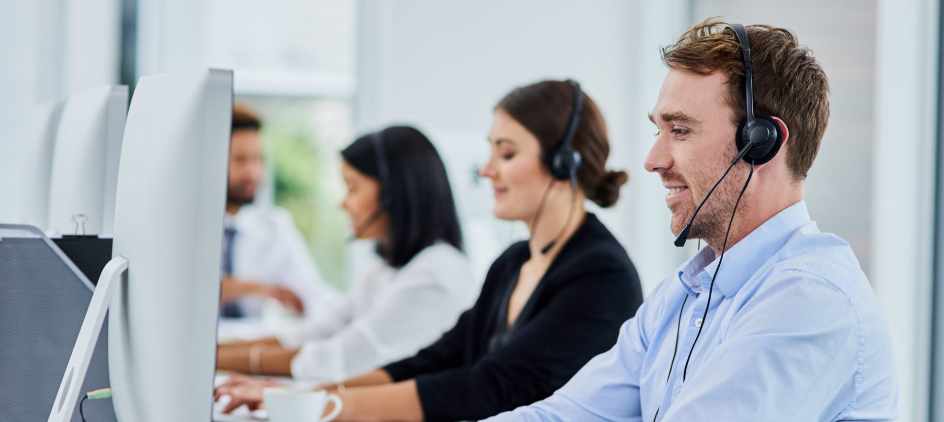 Smiling agent in a headset typing at a monitor on a busy support floor, answering bioprocessing customers