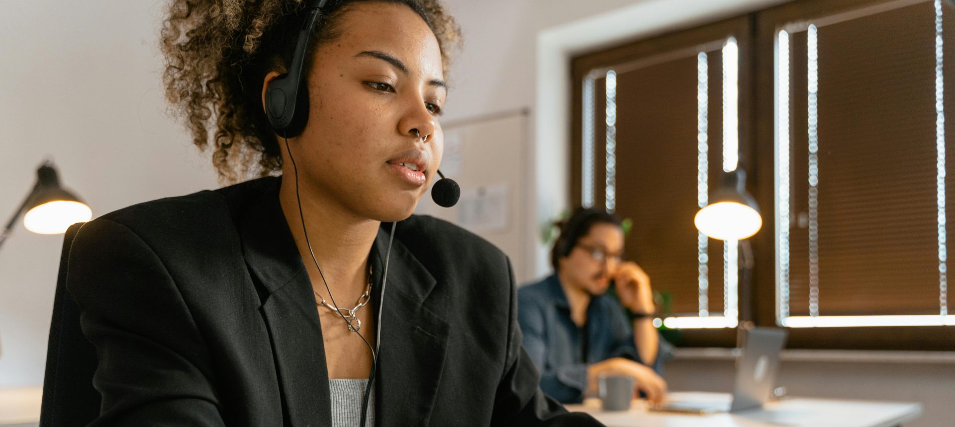 Contact center agent in a headset typing a case note while a colleague takes a call behind her