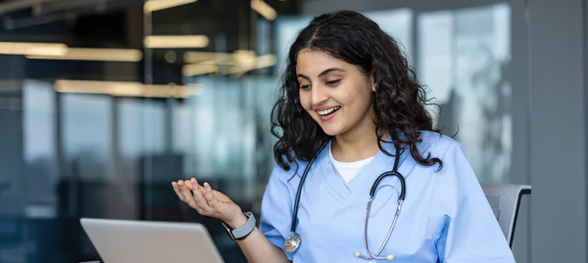 Smiling nurse in blue scrubs on a video call with a patient from her desk, a clipboard beside the laptop