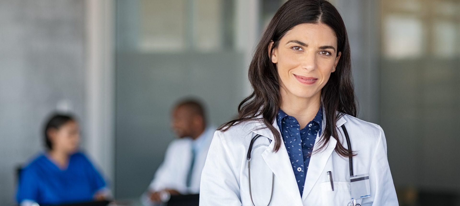 Smiling doctor in a white coat with a stethoscope using a tablet, with clinical colleagues meeting in the background, representing healthcare outsourcing in the Philippines