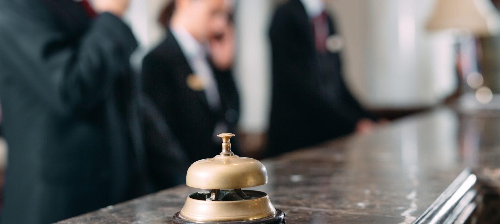 Brass service bell on a marble hotel counter with front-desk staff on the phone behind, the desk a PMS support team serves