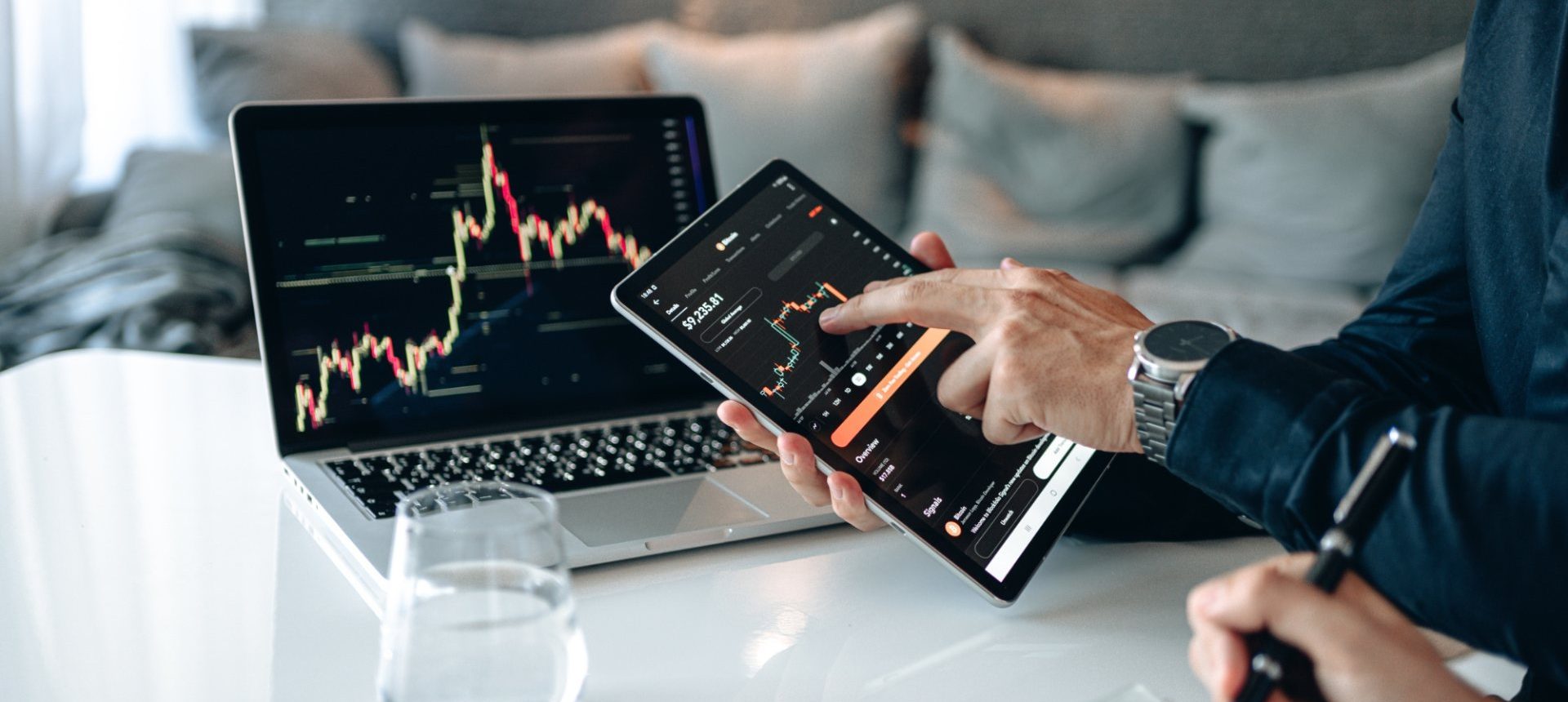 Hand pointing at a trading app on a tablet beside a laptop chart, the client view of an investment platform