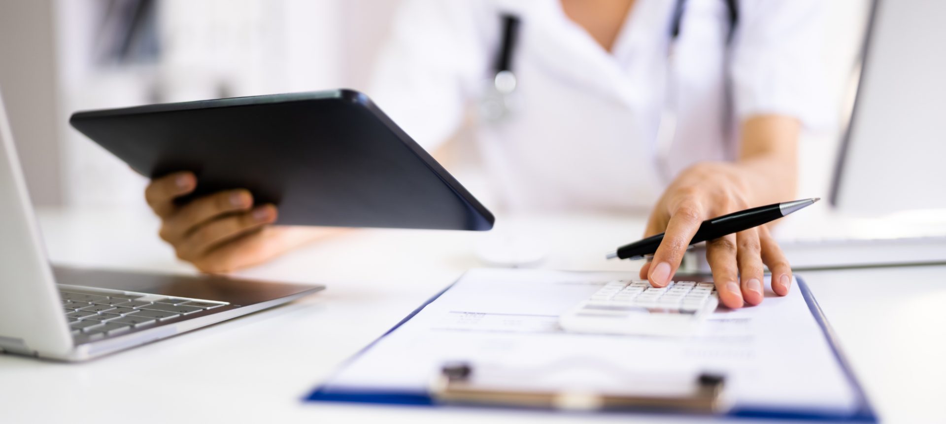 Clinician holding a tablet and tapping a calculator over a clipboard, checking the charges a laboratory billing team submits