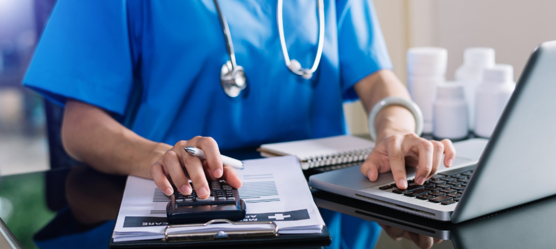 Medical billing specialist in scrubs calculating a patient claim on a clipboard beside a laptop