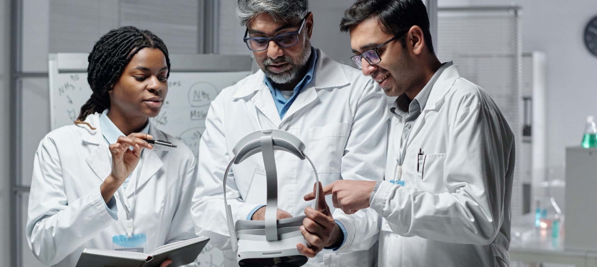Three scientists in lab coats examining a wearable headset and taking notes, the kind of product a medical device team backs