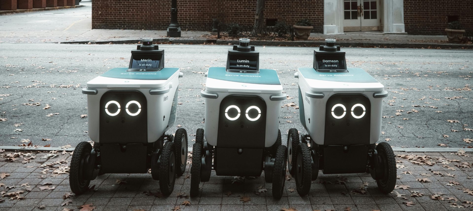 Three sidewalk delivery robots lined up on a city street, the fleet a last-mile robot operations team monitors