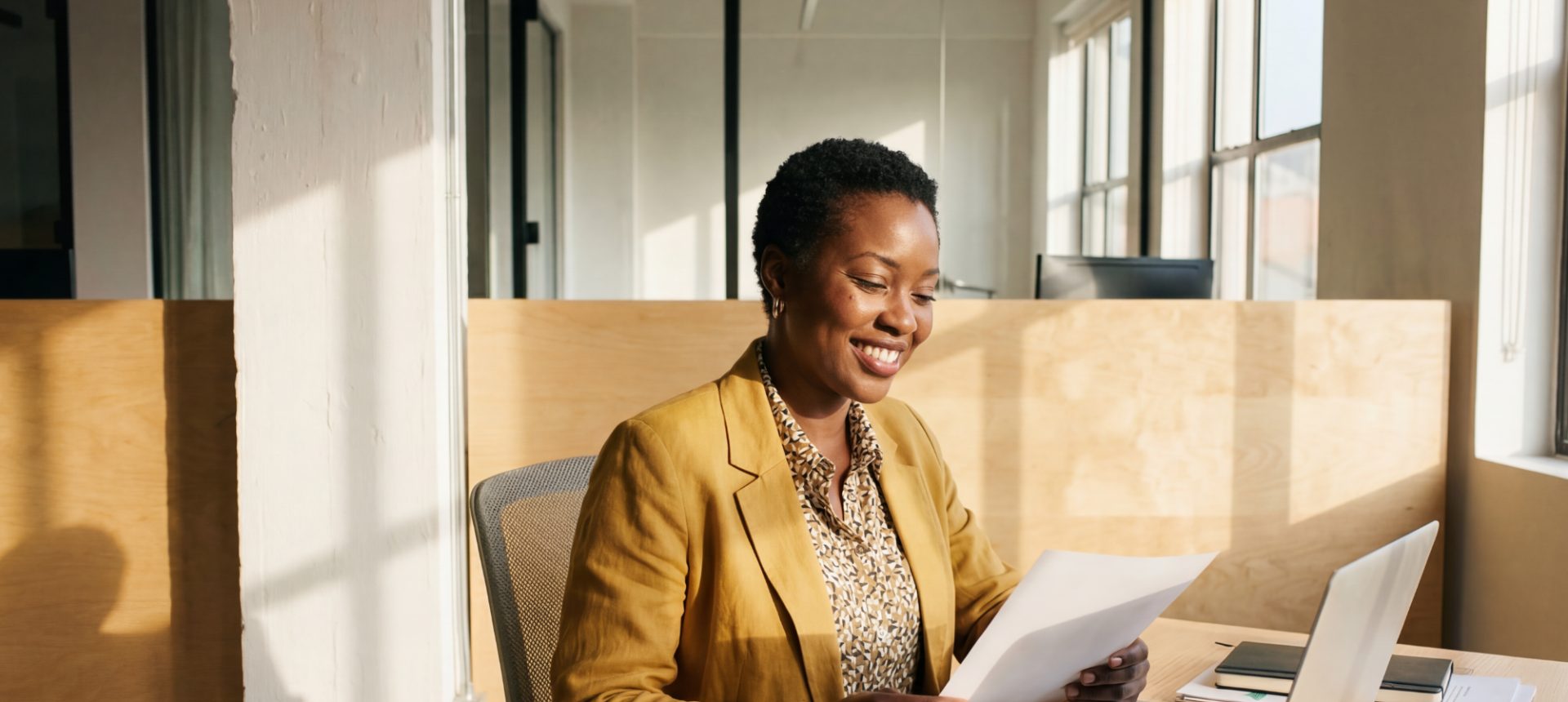 Smiling recovery specialist reading a document at a sunlit desk beside her laptop, working a subrogation file