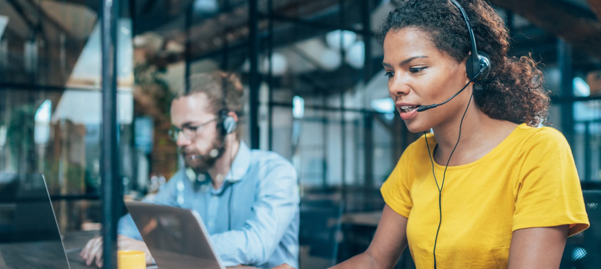 Support agent in a yellow top with a headset typing a reply on her laptop, a colleague working beside her