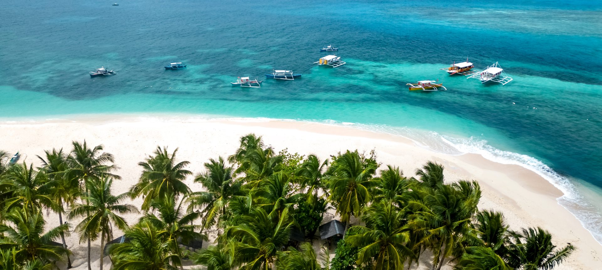 Aerial view of a white-sand Philippine beach lined with palm trees, with outrigger bangka boats on turquoise water, representing travel and OTA customer support outsourcing in the Philippines