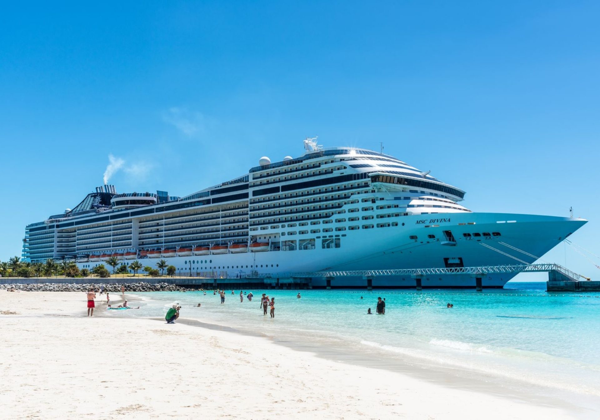 Cruise ship docked beside a white-sand beach with guests in the water, a port day cruise guest teams plan for