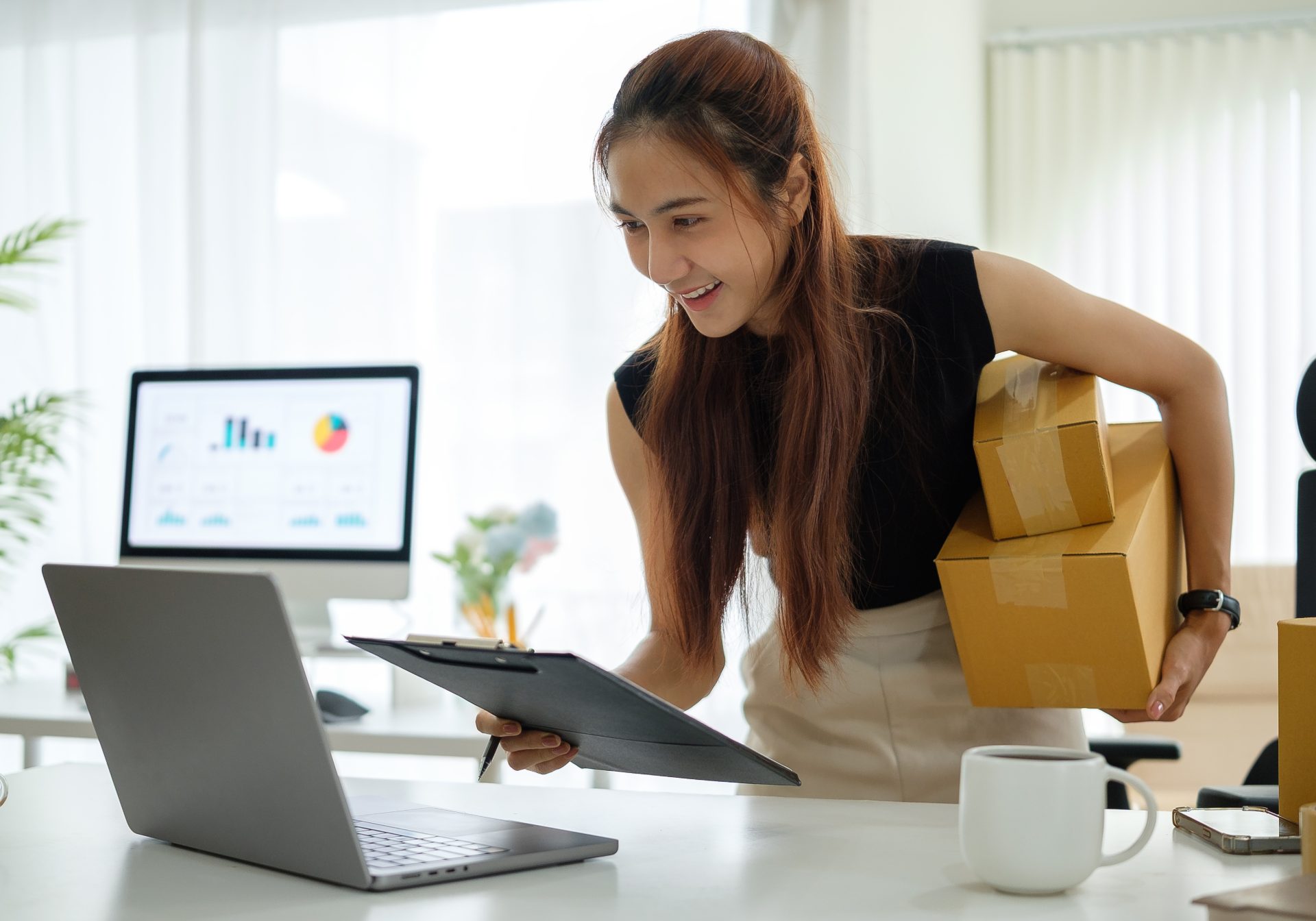 Seller checking an order list against her laptop while carrying parcels, the fulfillment a dropshipping team tracks