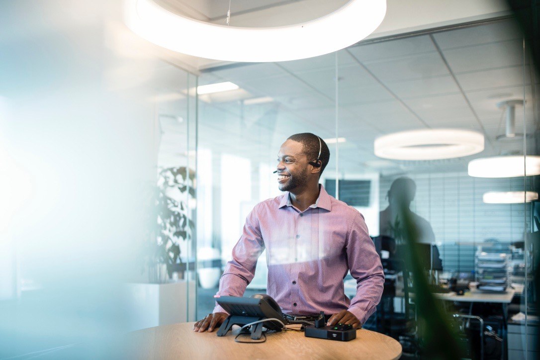 Smiling call center agent with a headset standing at a desk phone in a glass-walled contact center office