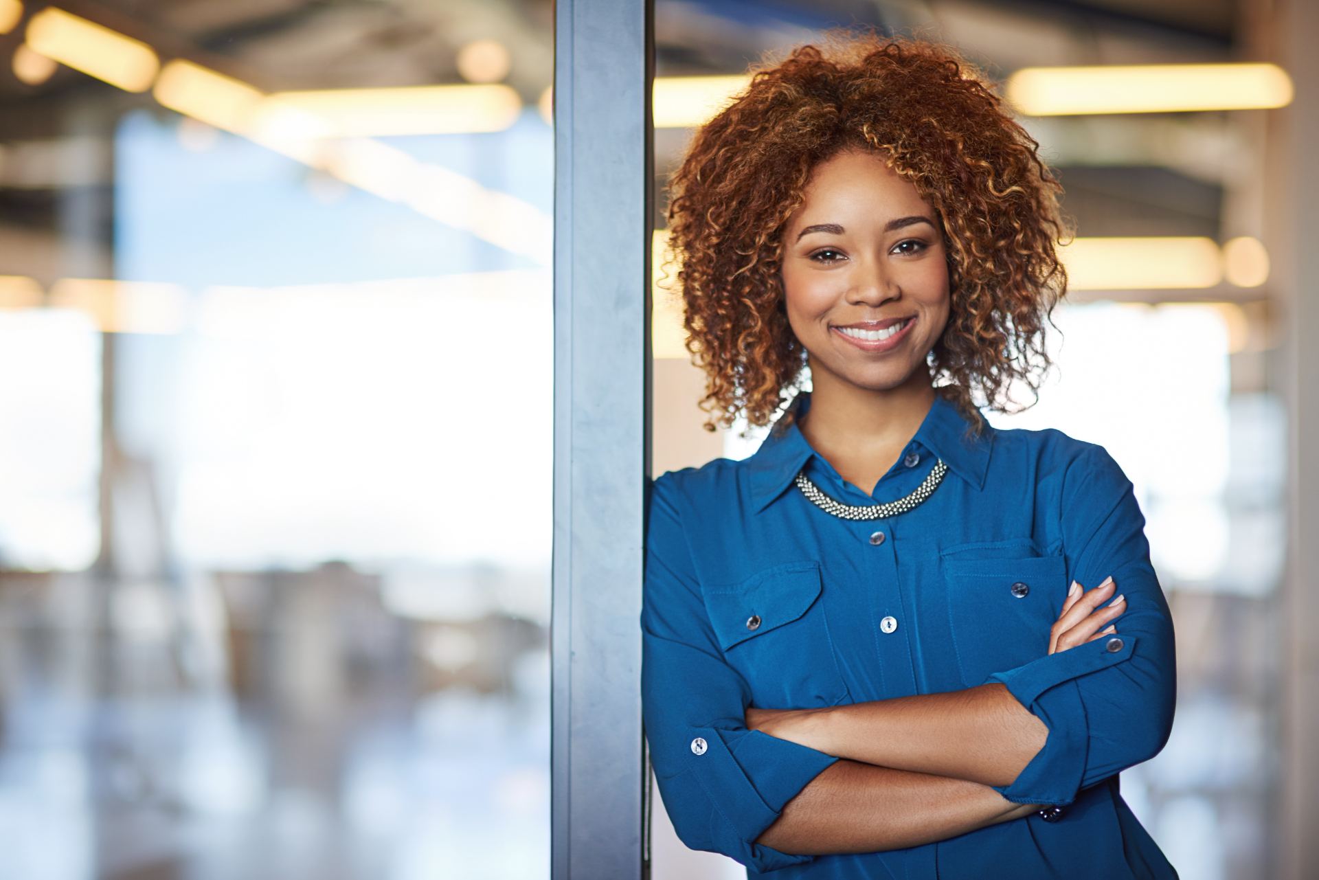 Smiling business professional with arms crossed, leaning against a glass wall in a modern office