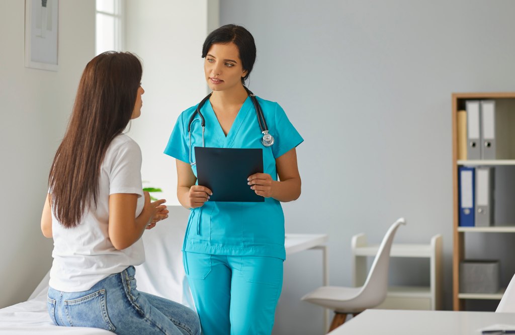 Nurse holding a clipboard talking with a patient in an exam room, representing health plan and payer member services from the Philippines