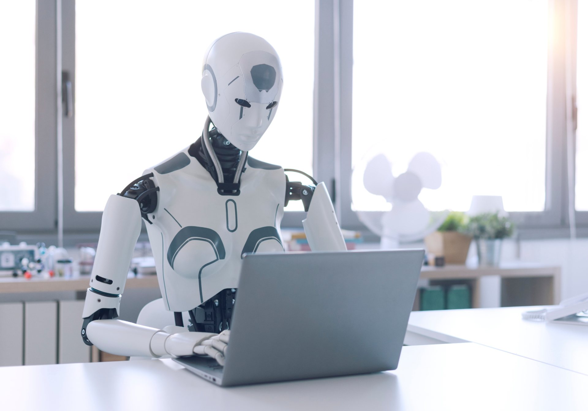 Humanoid robot typing on a laptop at an office desk, the kind of task a teleoperator helps it finish