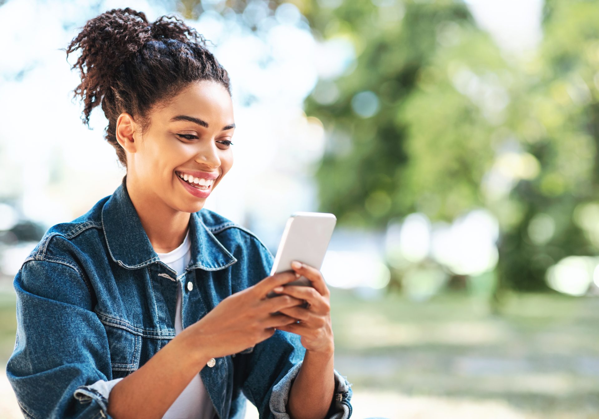 Smiling young woman in a denim jacket reading a support reply on her phone outdoors