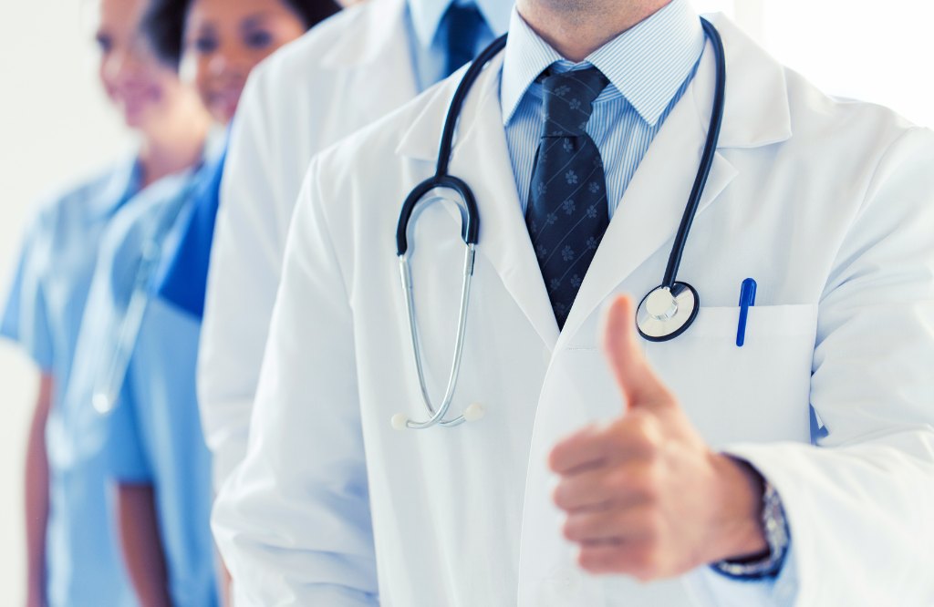 Physician in a white coat and stethoscope giving a thumbs-up in front of his care team, representing medical credentialing and provider enrollment from the Philippines