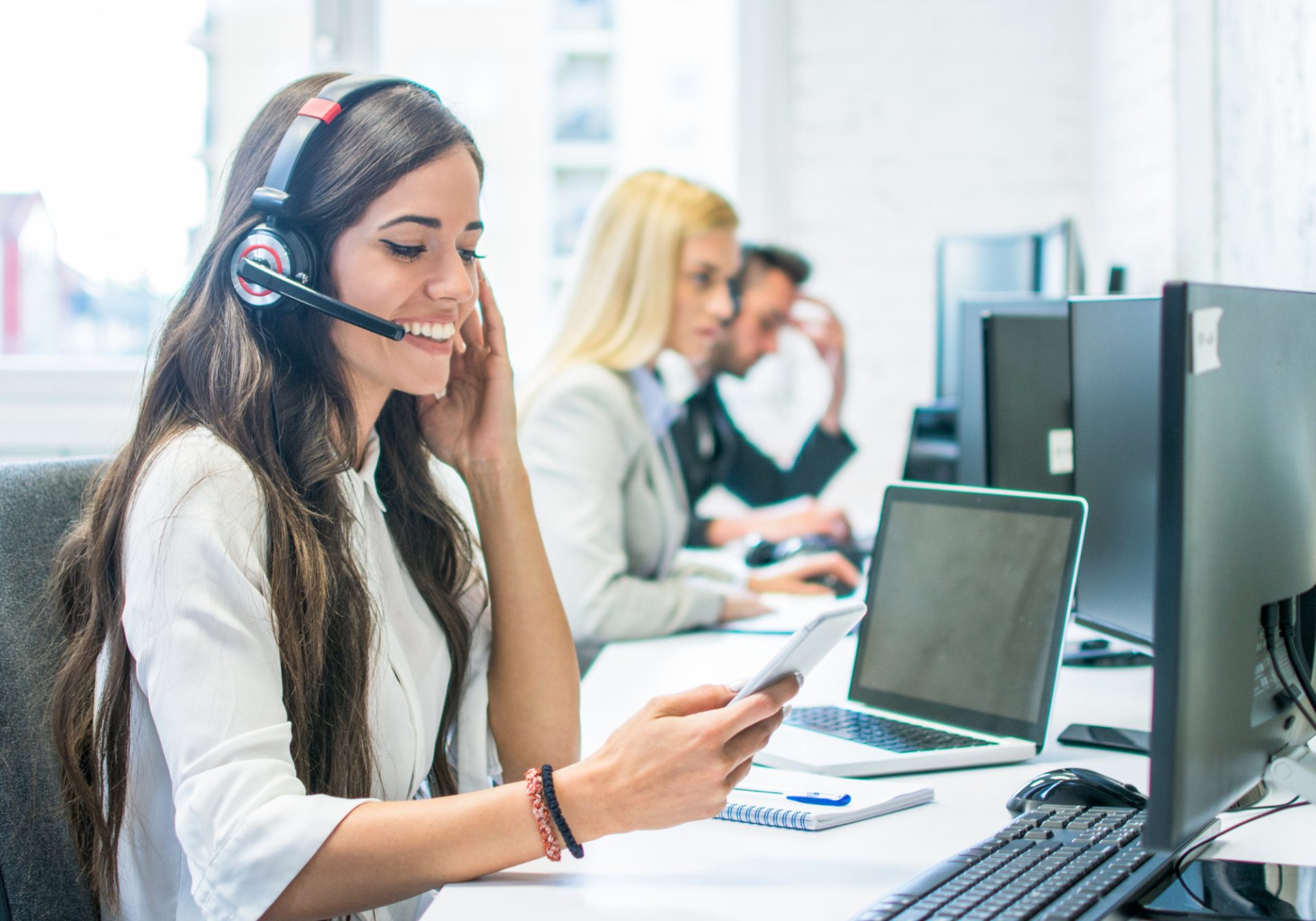 Smiling retail support agent with a headset checking a shopper’s order on her phone at a busy support desk