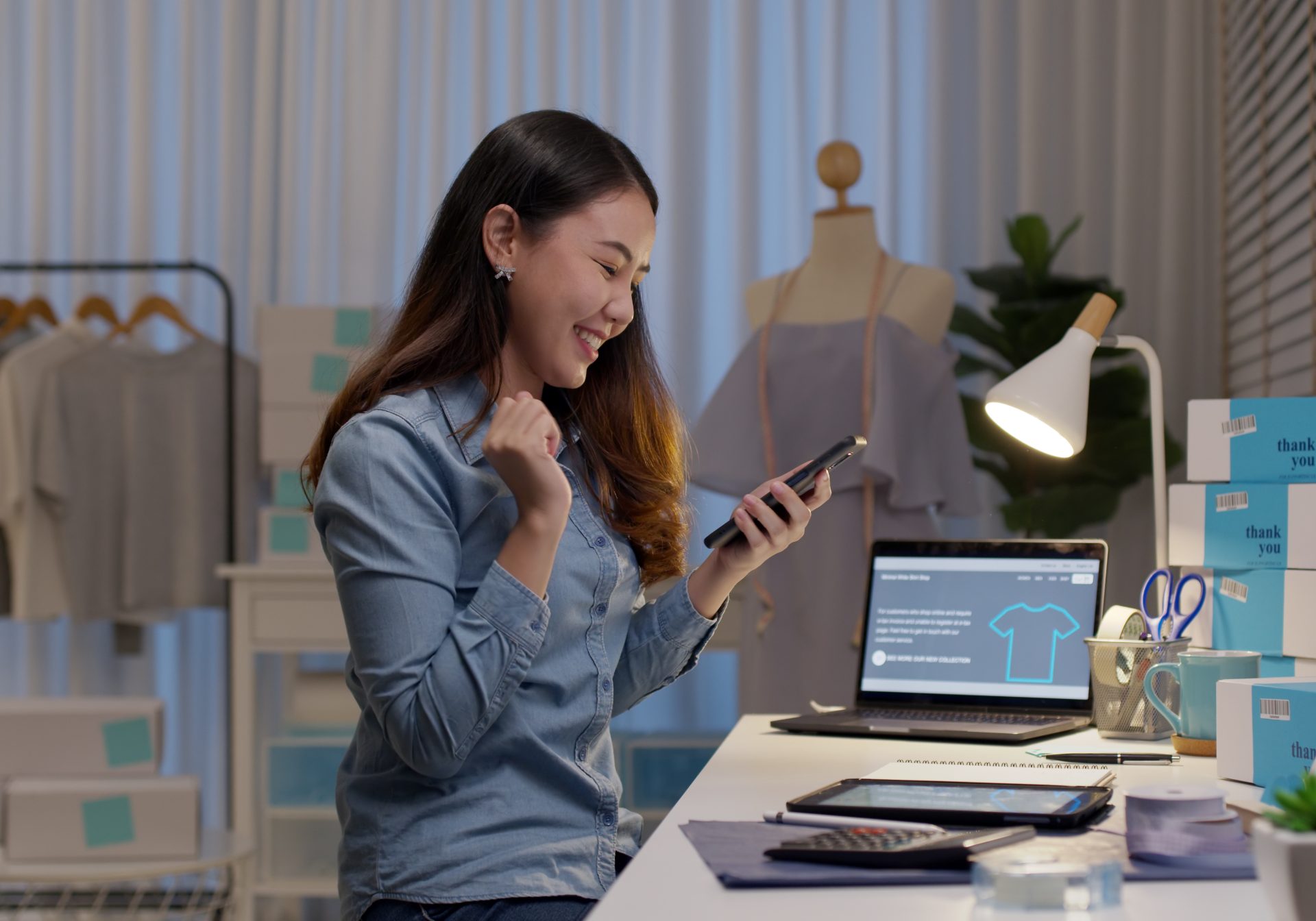 Online store owner celebrating an order on her phone beside her storefront laptop and packed boxes