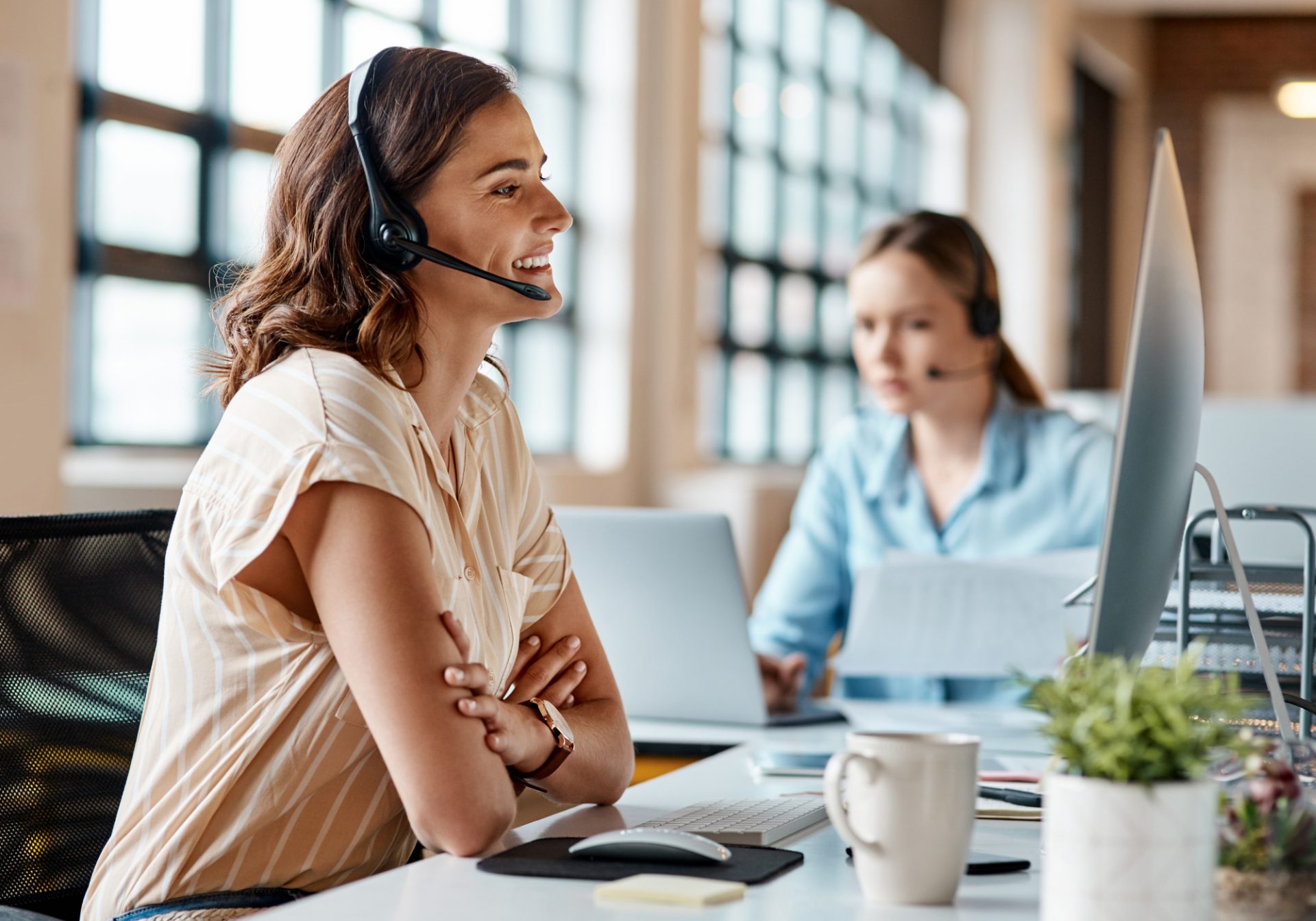 Smiling guest services agent with a headset listening to a traveler at her desk, a colleague working behind her
