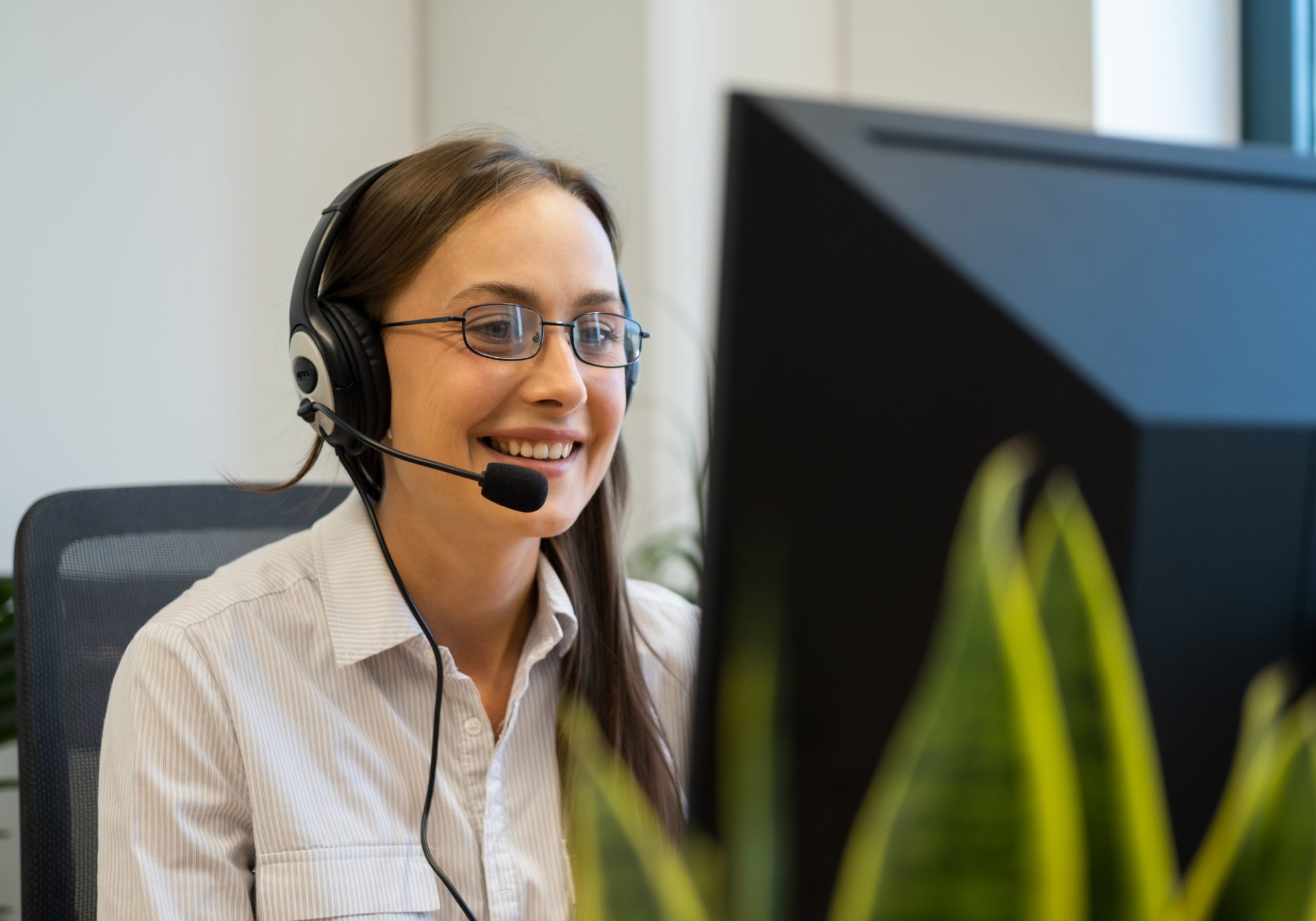 Smiling utilities care agent with glasses and a headset explaining a bill to a customer at her computer