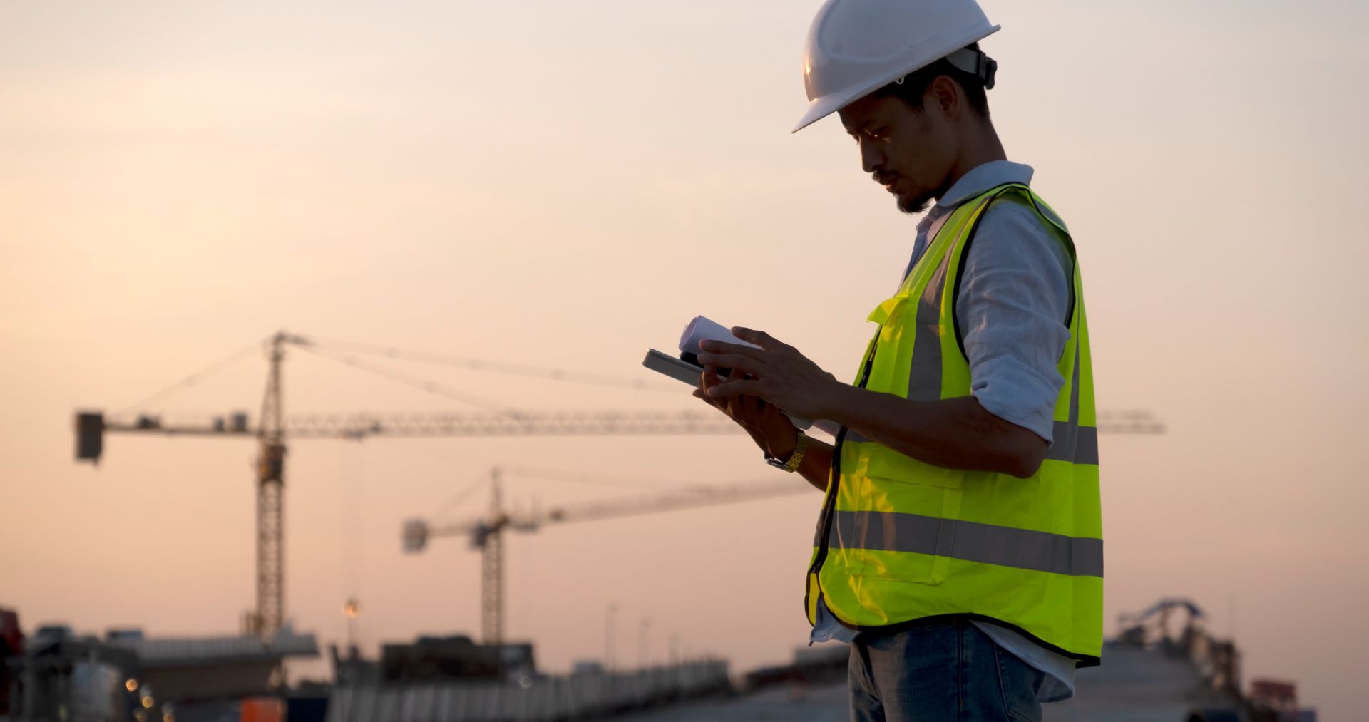 Site engineer in a hard hat checking a tablet near tower cranes at dusk, for a post on utility arrears payment plan